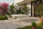 A white metal outdoor dining set consisting of a rectangular table and six matching chairs, arranged on a light stone patio in a modern backyard setting with lush green plants and a pink flowering bush.